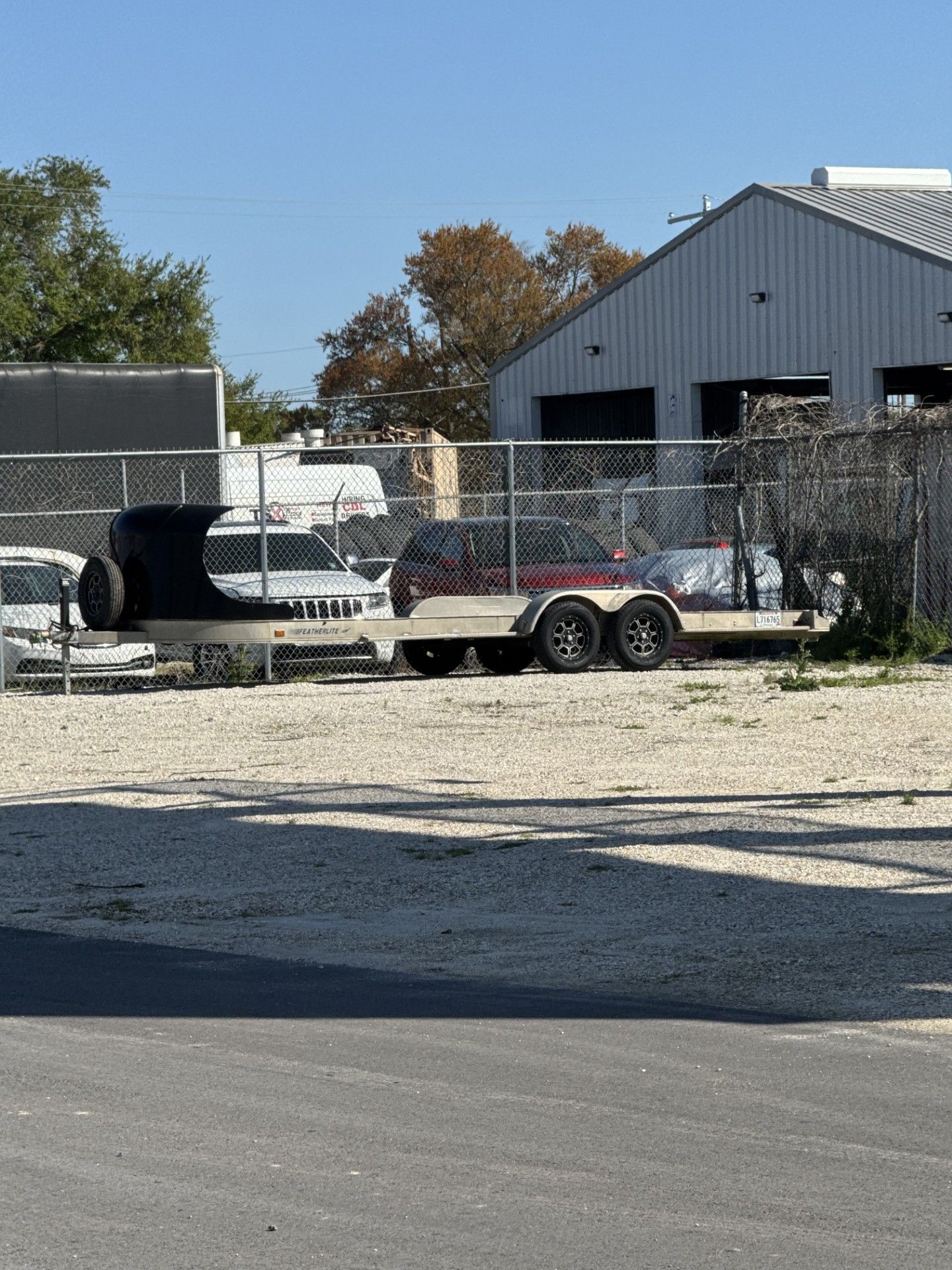 An empty tandem-axle trailer with a black front guard parked on a gravel lot next to a chain-link fence and a building.