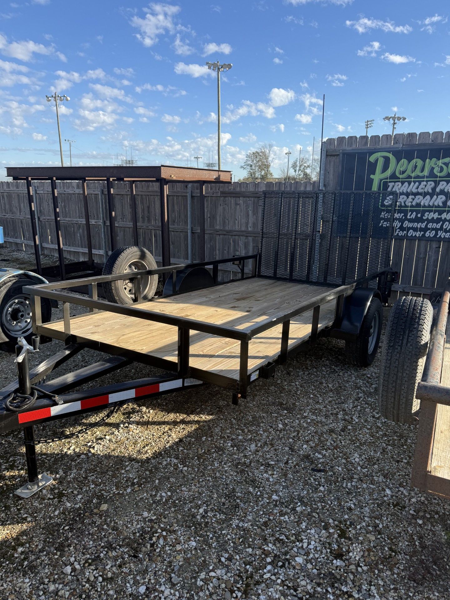 A black utility trailer with a wooden bed parked on gravel in front of a fence and a business sign.
