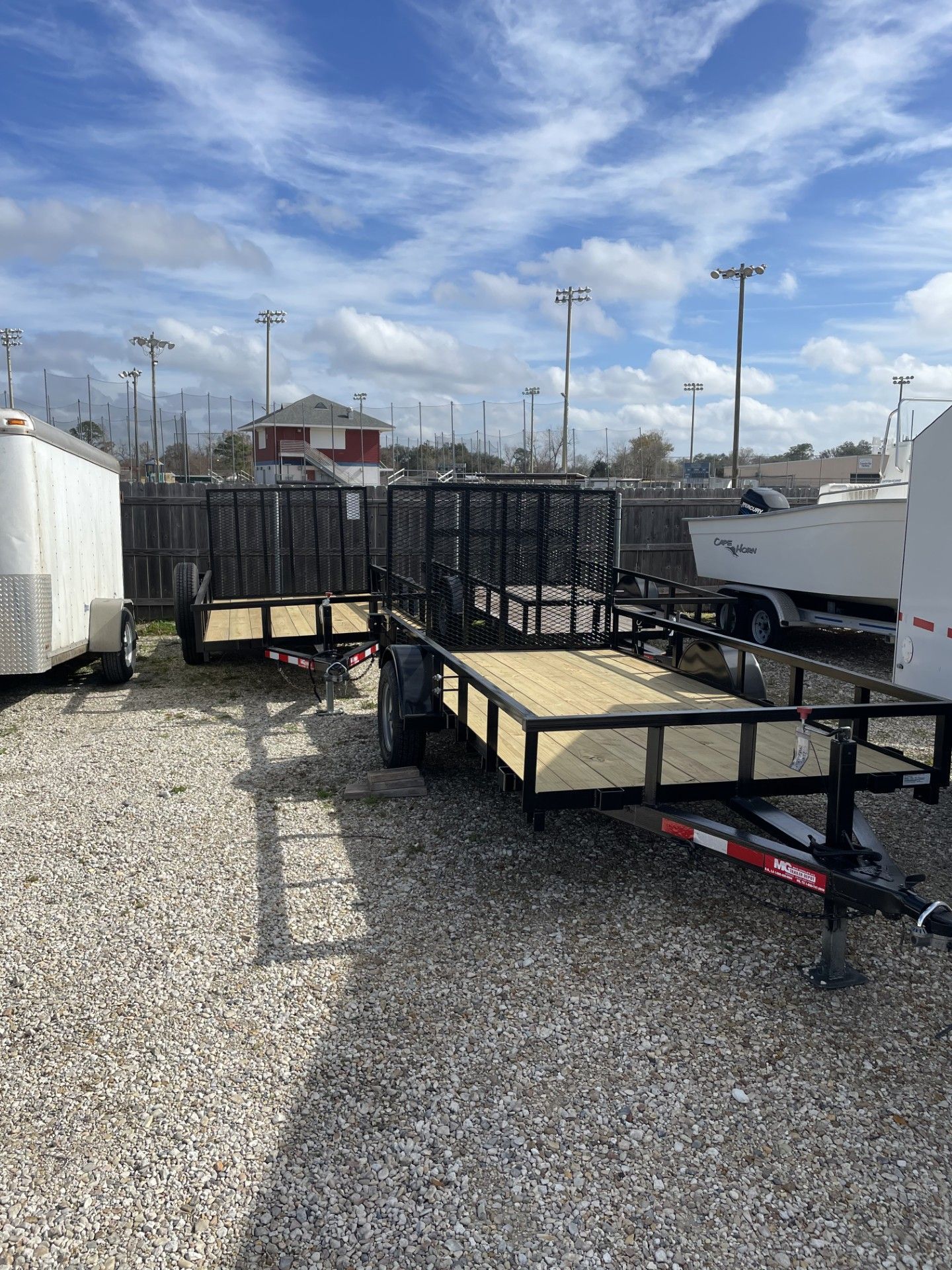 A gravel lot with utility trailers, including one with a wooden deck and black metal frame, under a partly cloudy sky.