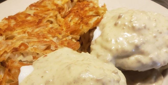 A breakfast plate featuring crispy golden-brown hash browns and biscuits covered in thick, white sausage gravy.