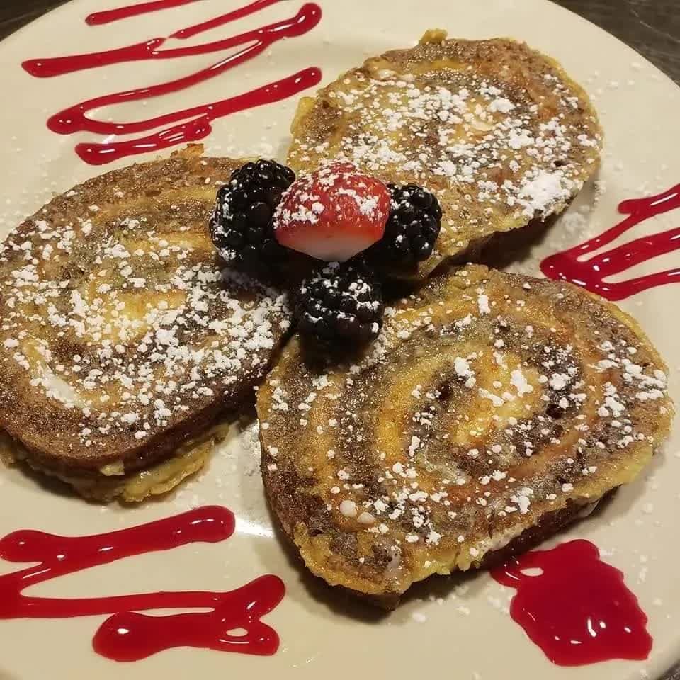 Three cinnamon roll-style French toast slices topped with powdered sugar, blackberries, and strawberry, with berry glaze.