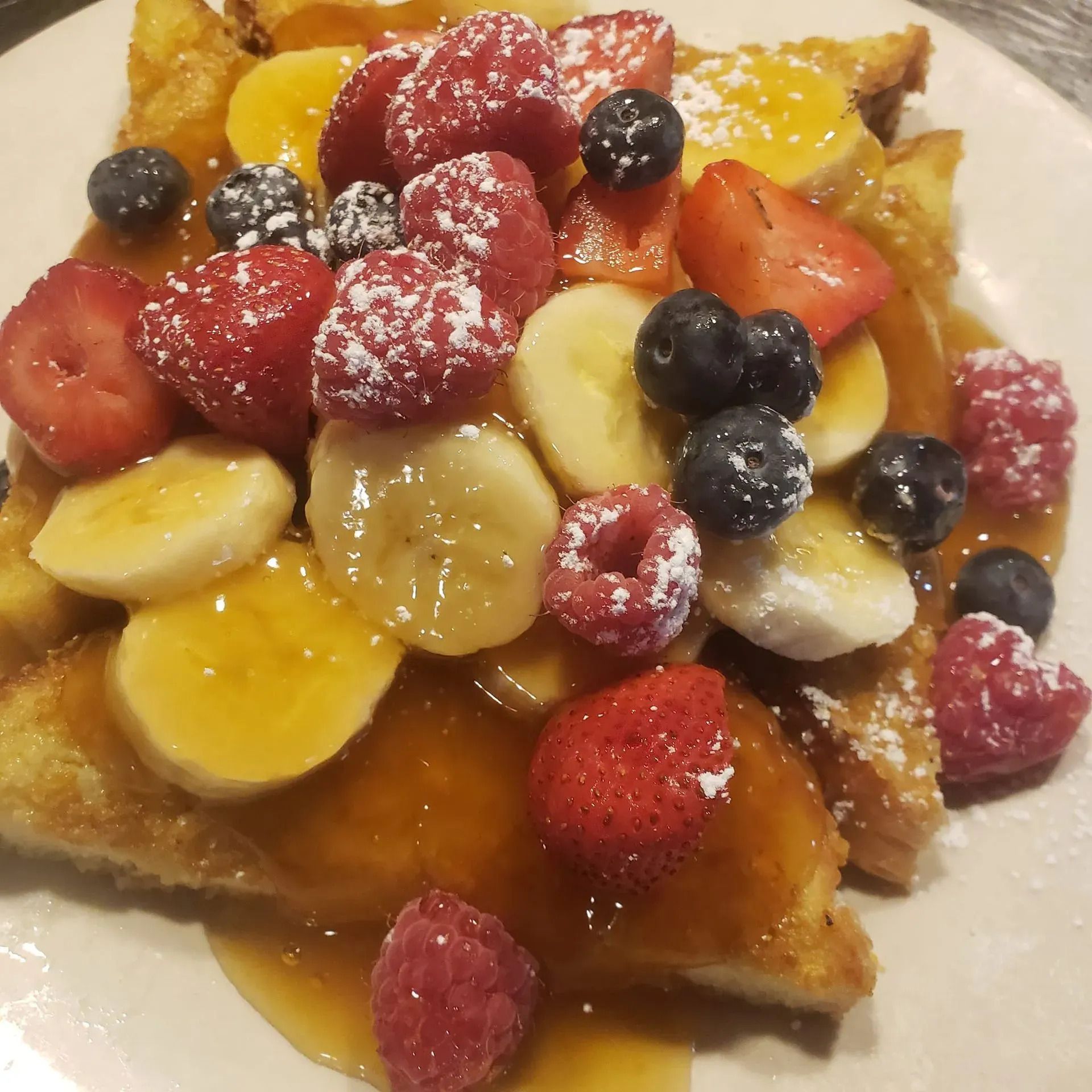 French toast topped with fresh strawberries, raspberries, blueberries, banana slices, and syrup, dusted with sugar.