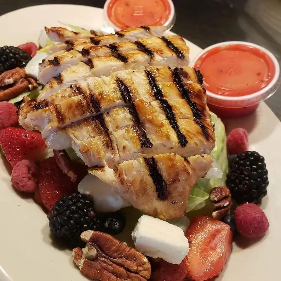 A grilled chicken salad on a white plate with strawberries, blackberries, pecans, feta cheese, and two cups of dressing.