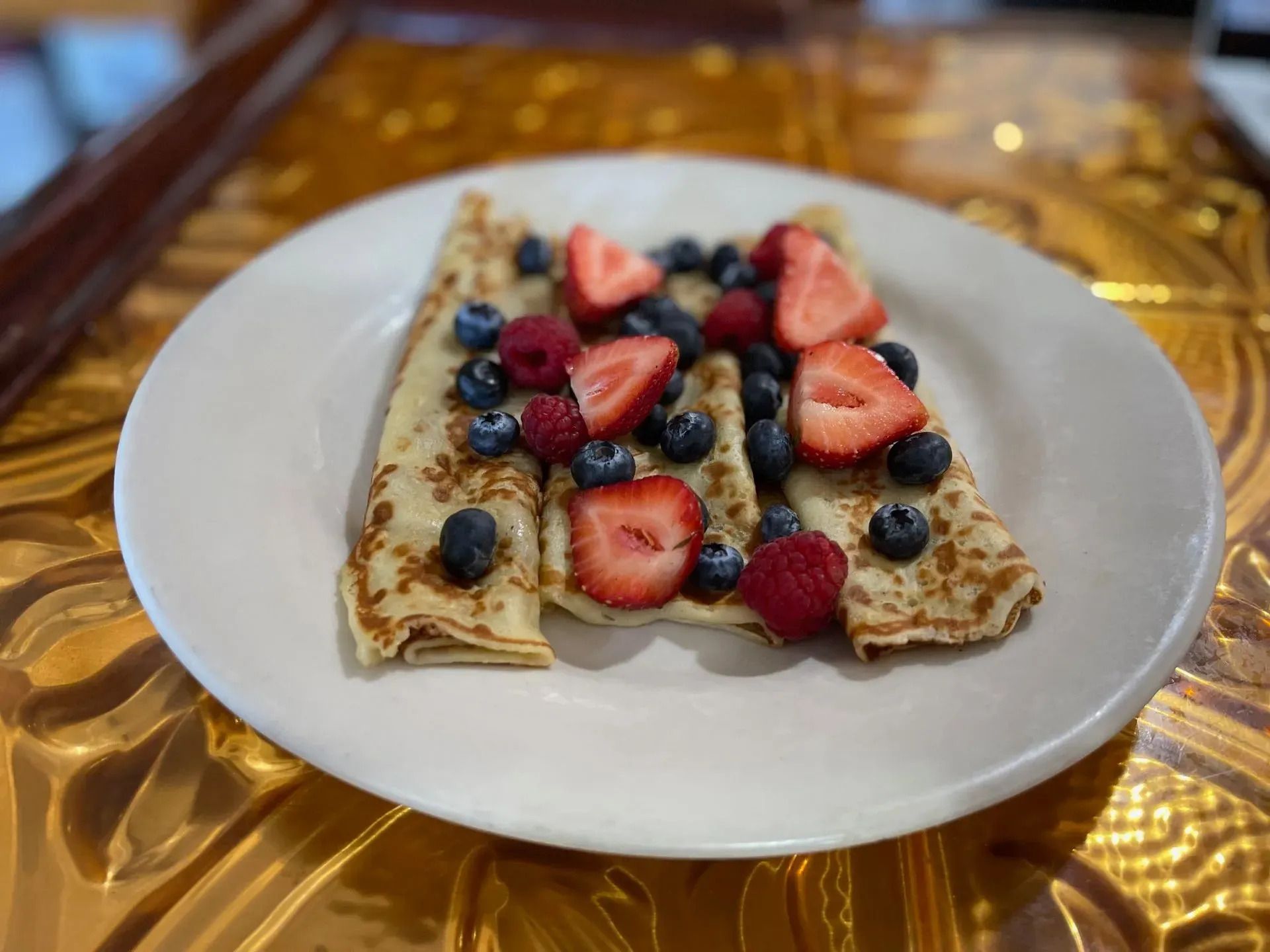 Three thin crepes topped with fresh strawberries, blueberries, and raspberries on a white plate over a gold surface.