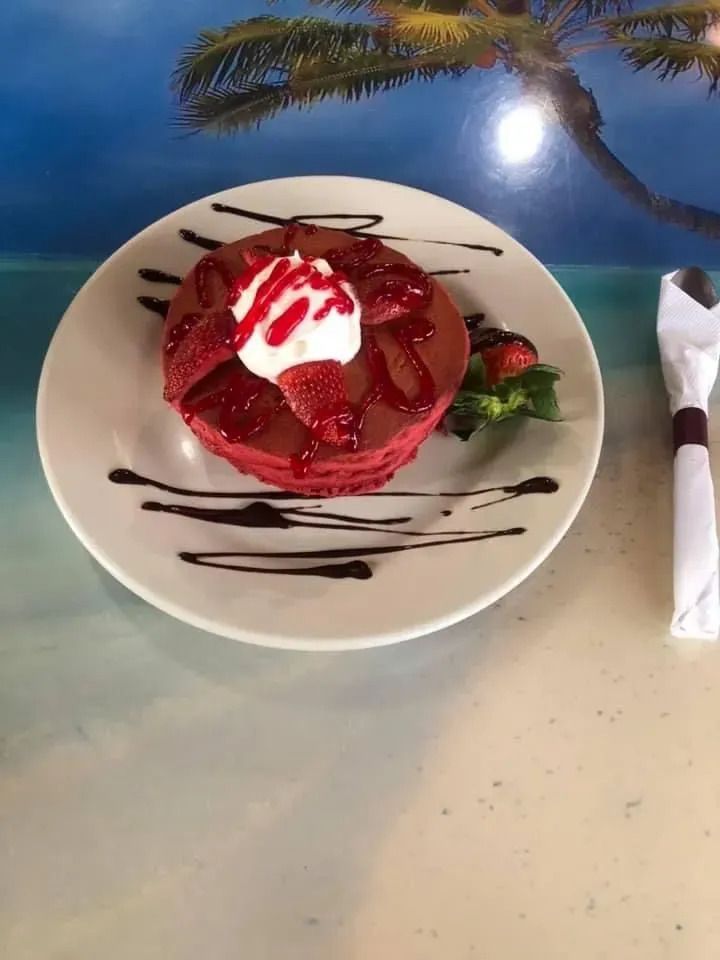 A stack of red velvet pancakes topped with whipped cream, strawberry syrup, and sliced strawberries, served on a plate.