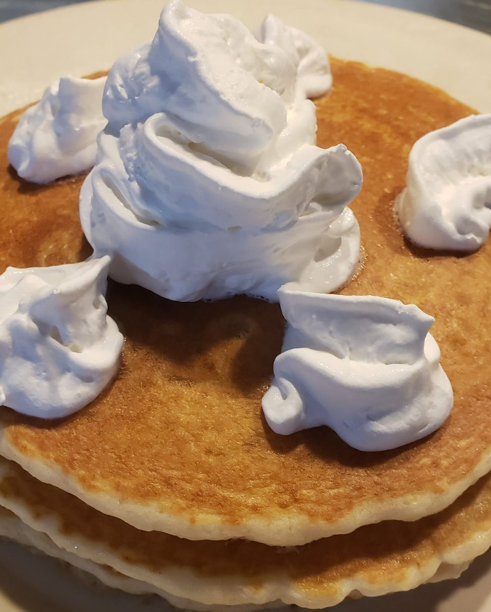 A stack of golden-brown pancakes topped with a generous swirl of whipped cream and several smaller dollops.