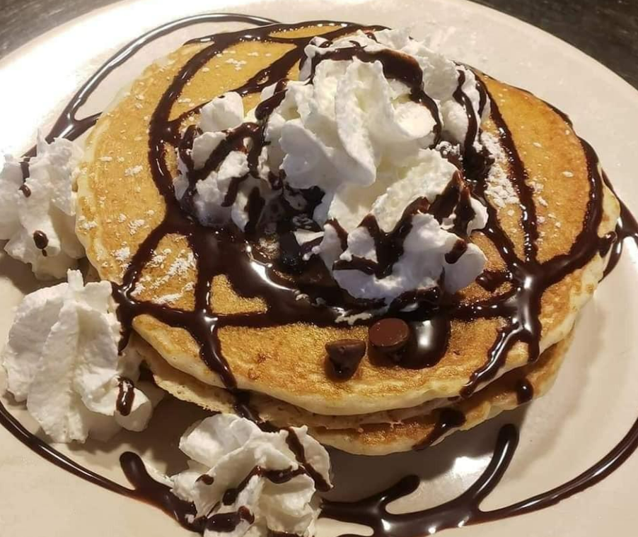 A stack of pancakes topped with whipped cream, chocolate chips, and a drizzle of chocolate sauce on a white plate.