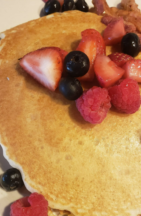 A golden pancake topped with fresh strawberries, blueberries, raspberries, and strips of crispy bacon.