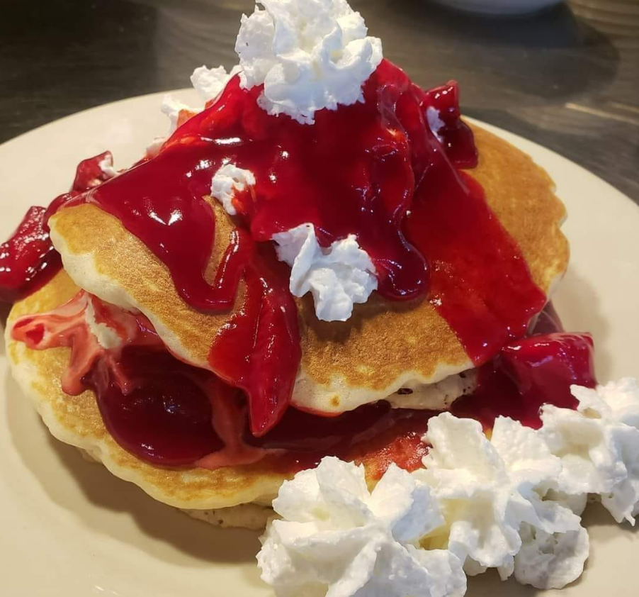 A stack of golden pancakes topped with bright red berry sauce and swirls of white whipped cream on a cream-colored plate.