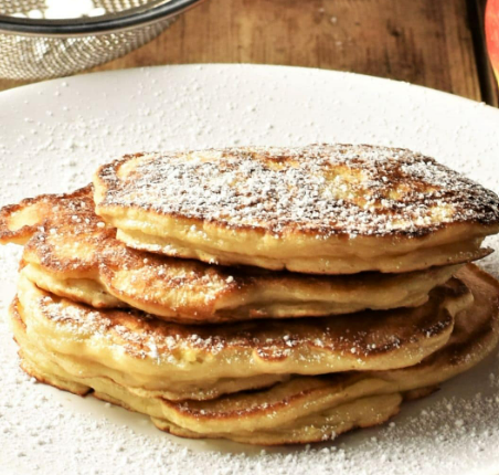 A stack of golden-brown pancakes on a white plate, dusted with powdered sugar against a wooden background.