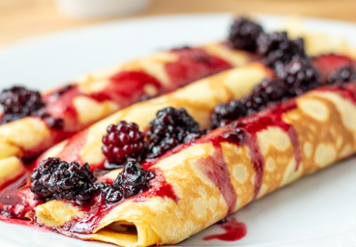 Two rolled crepes on a white plate, topped with fresh blackberries and drizzled with a vibrant red berry sauce.