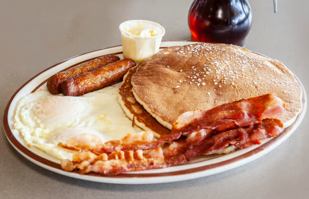 A breakfast plate with two eggs, two sausages, several strips of bacon, pancakes, and a small cup of butter.