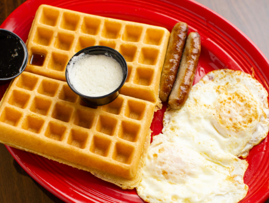 A breakfast plate with a waffle, two sausages, two fried eggs, and small containers of butter and syrup on a red plate.