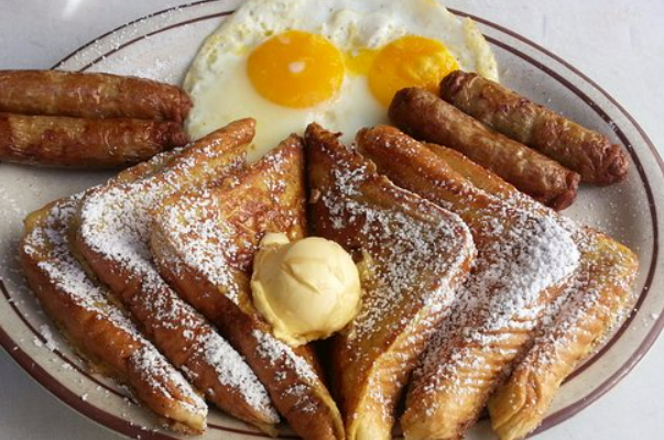 A breakfast plate with French toast sprinkled with powdered sugar, a scoop of butter, two sausage links, and two eggs.