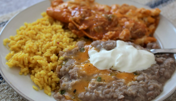 A plate of Mexican-style food featuring yellow rice, refried beans topped with sour cream, and an enchilada with sauce.
