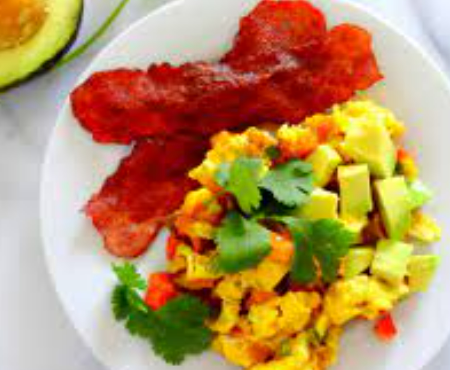 A breakfast plate featuring scrambled eggs with avocado and cilantro, served with two strips of crispy bacon.