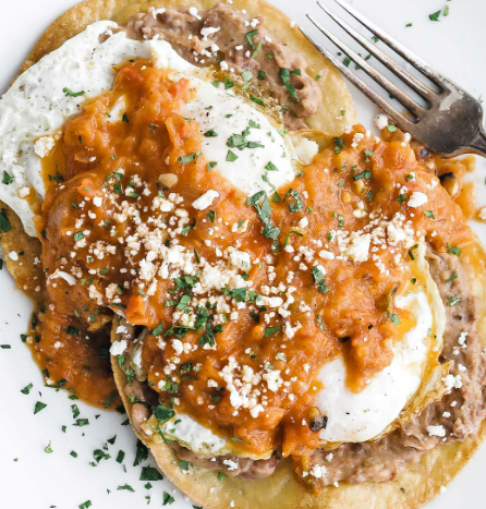 Huevos rancheros with fried eggs on tortillas topped with beans, red salsa, queso fresco, and cilantro on a white plate.