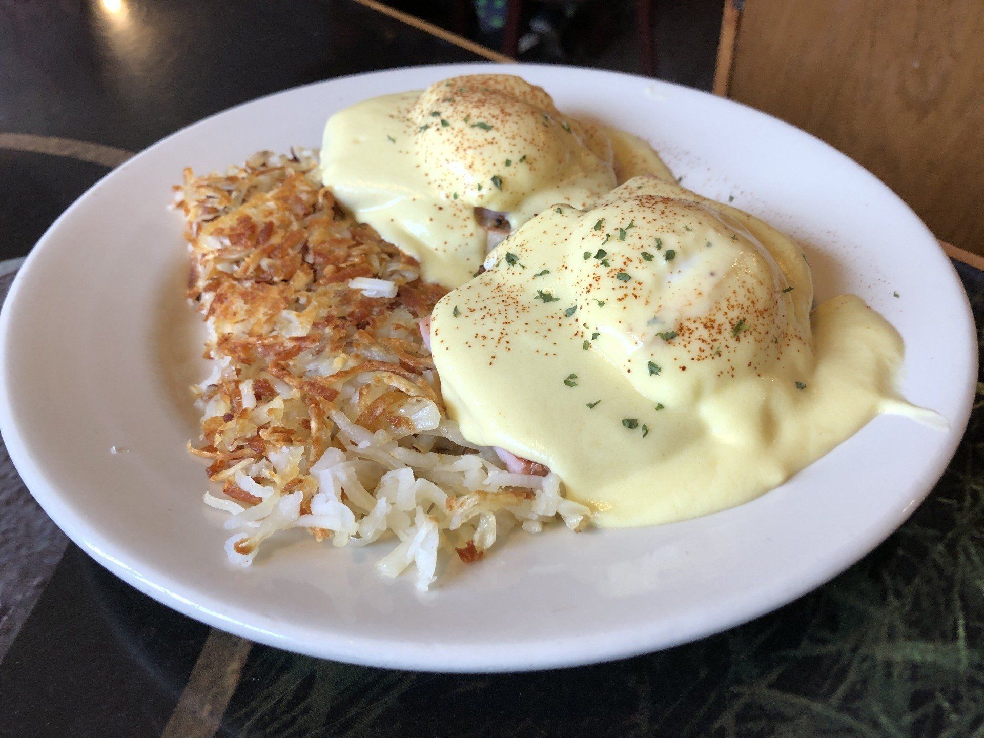 Two eggs Benedict topped with hollandaise sauce and herbs, served with a side of golden-brown shredded hash browns.