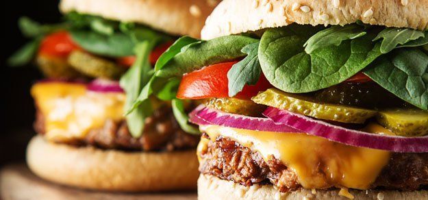 Two cheeseburgers stacked with fresh greens, tomato slices, red onion, and pickles on toasted buns.