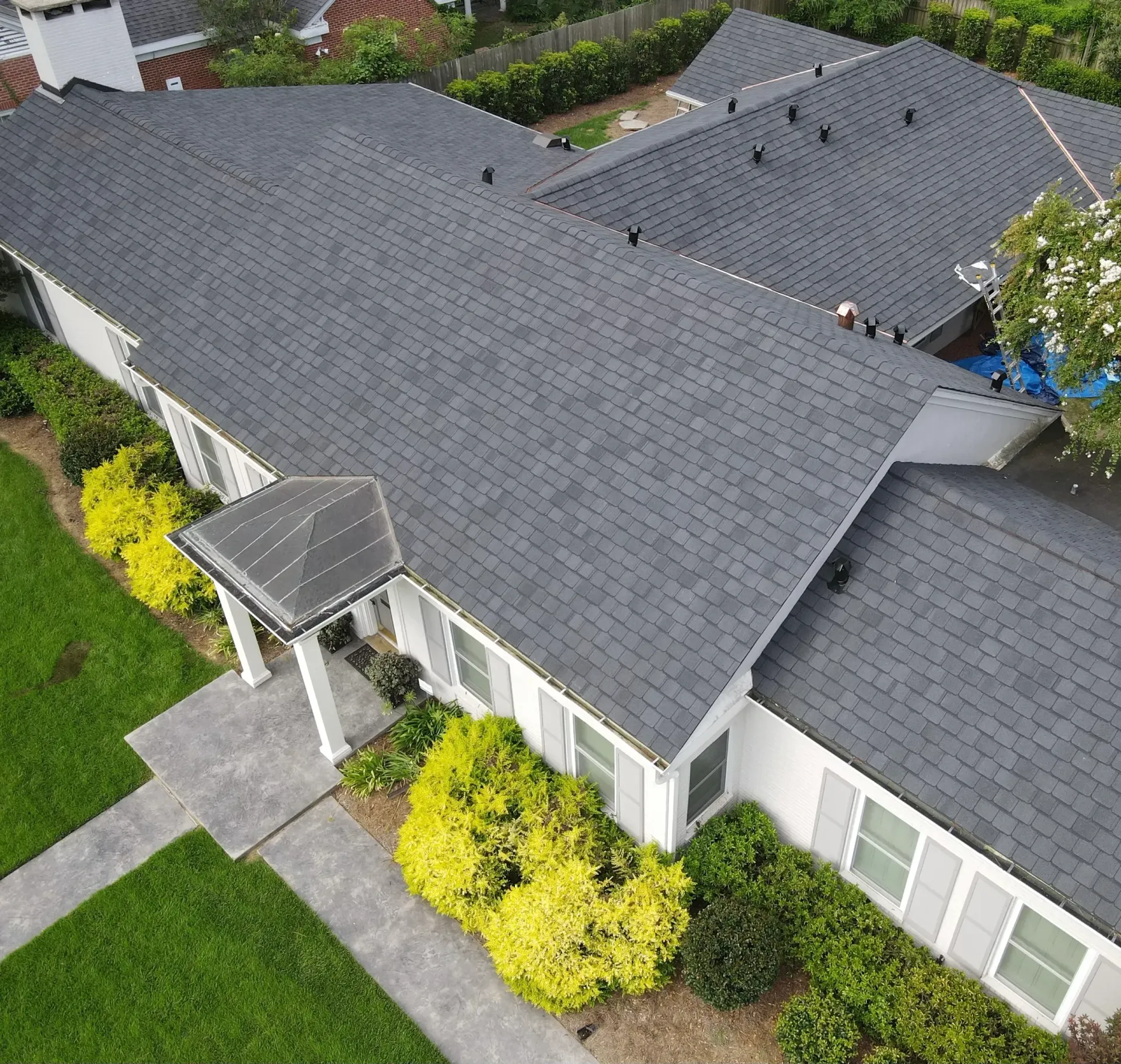Aerial view of a single-story house with a dark gray roof, front walkway, and green shrubs