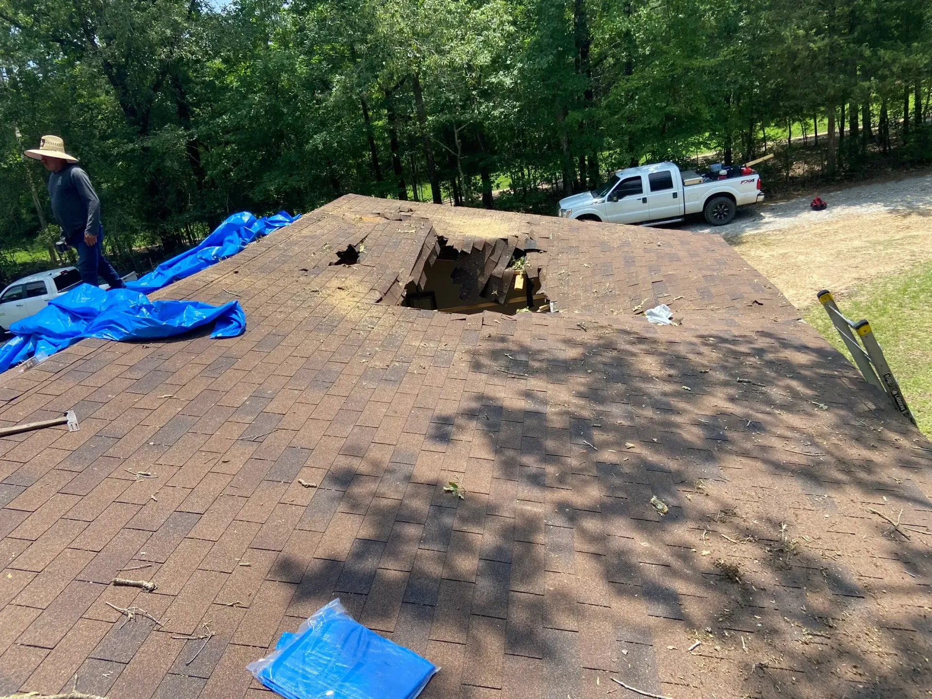 Roof under construction with blue tarps, a worker, and a white pickup truck in a wooded yard