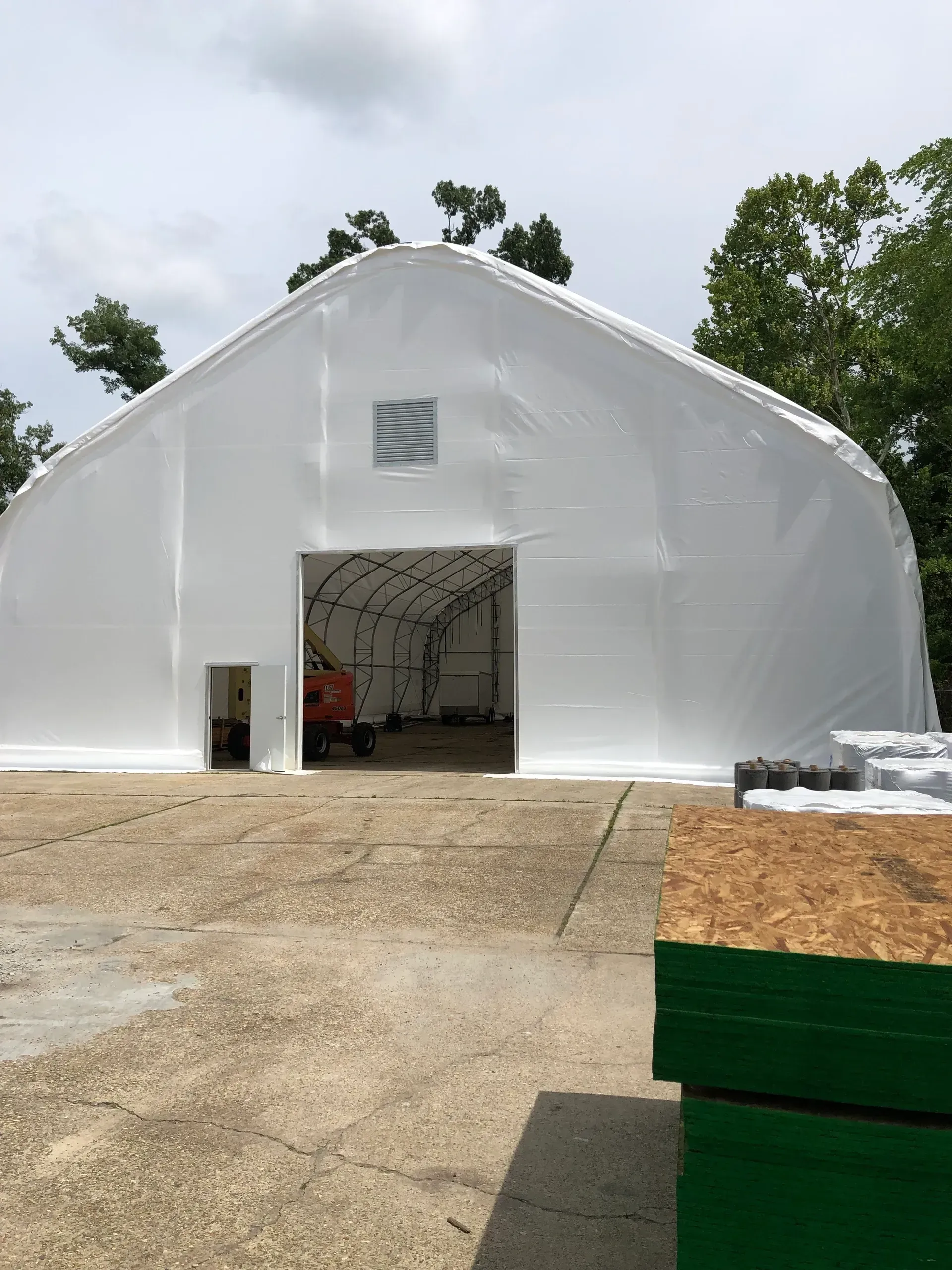 White tent-like storage building with an open doorway on a gravel lot, with trees in the background