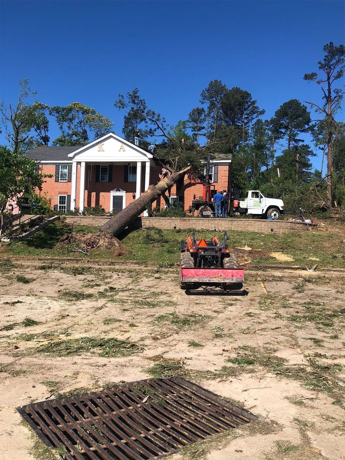 Brick house with a fallen tree, workers, and a white pickup truck in a yard under a clear blue sky