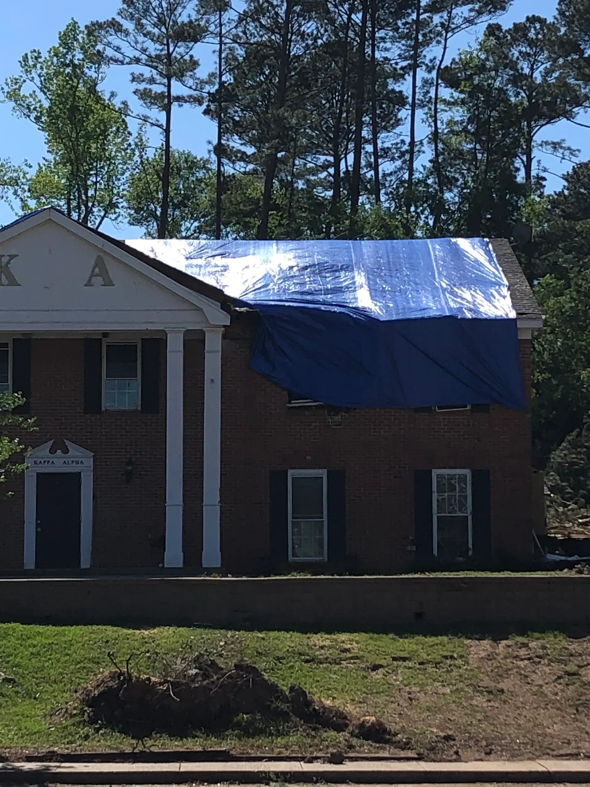 Two-story brick house with a blue tarp covering part of the roof after storm damage.