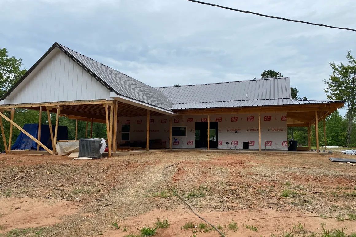 Partially built rural house with a metal roof and open porch on a dirt lot under cloudy sky