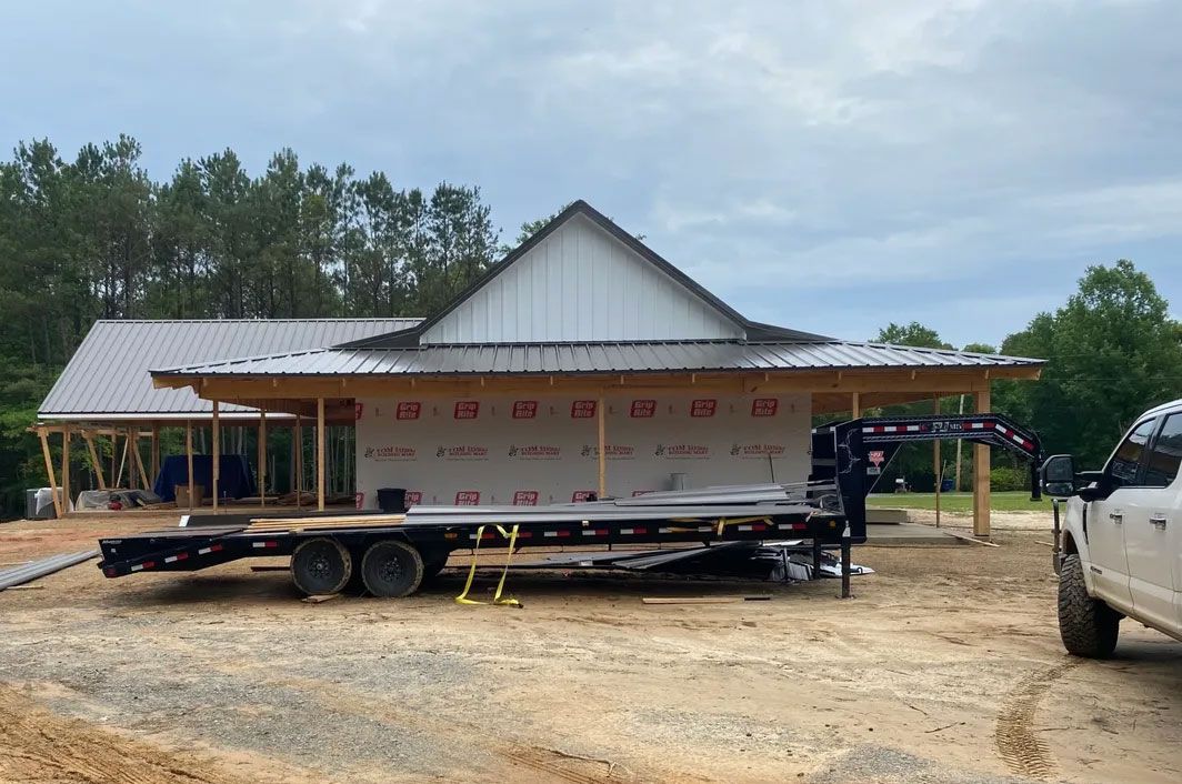 House under construction on a trailer in a dirt lot, with a pickup truck and trees nearby