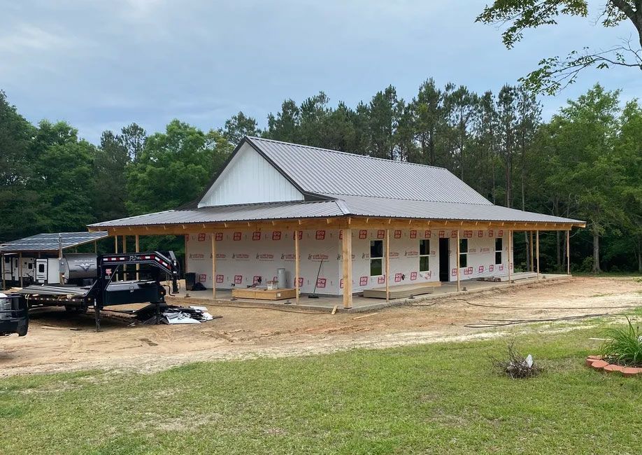 Partially built white house with metal roof, surrounded by dirt, grass, and construction materials under cloudy sky