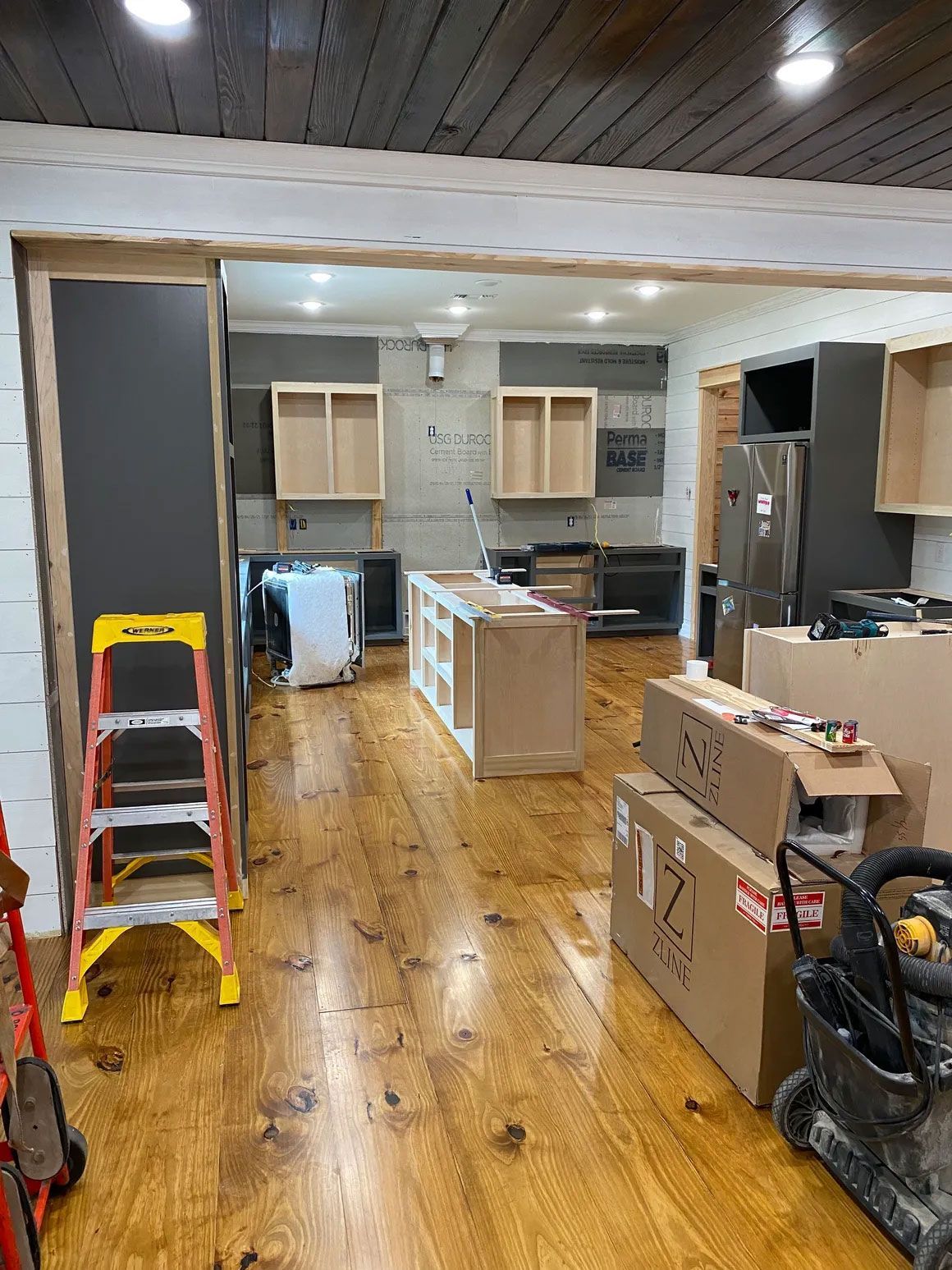 Kitchen under renovation with cabinets, appliances, boxes, and a yellow ladder on a wood floor.