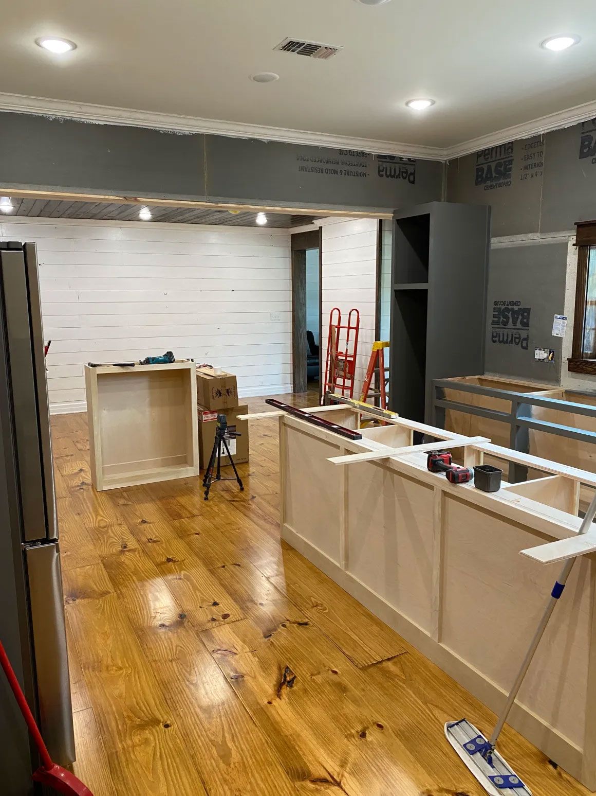 Kitchen under renovation with unfinished cabinets, wood floor, and construction materials scattered around.