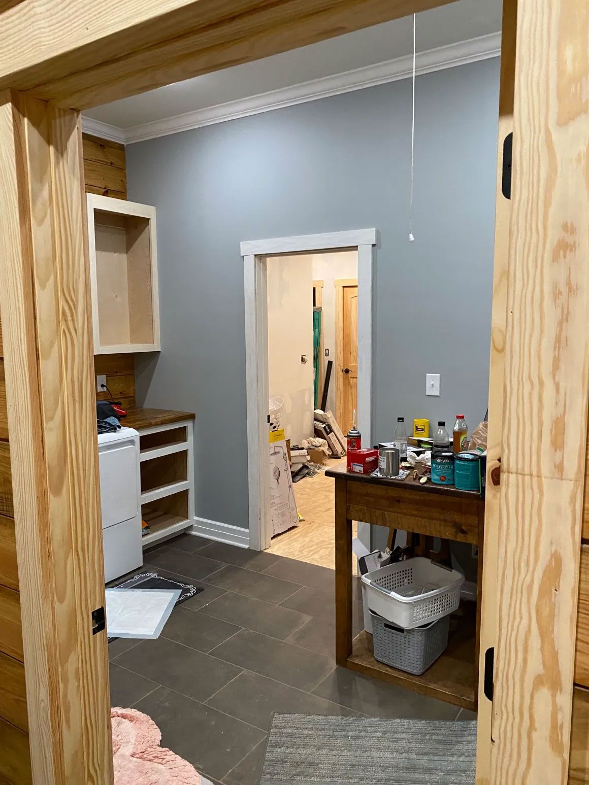 Small workshop room with gray walls, shelves, a workbench, and an open doorway to another room.