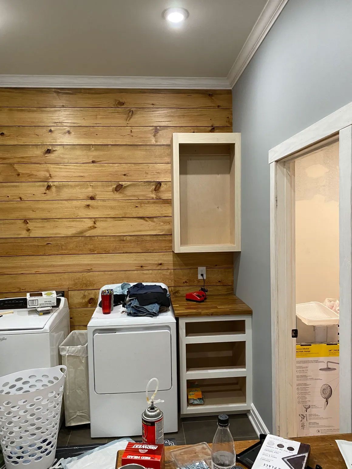Small room with wood accent wall, white cabinets, sink, laundry hamper, and an open doorway.