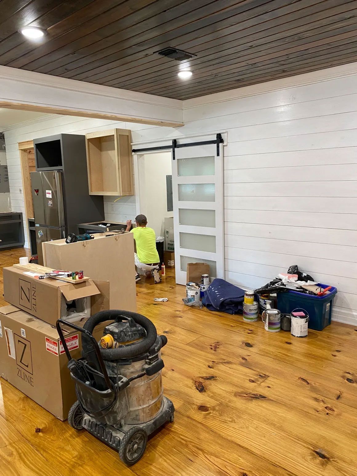 Renovation room with tools, boxes, and a worker kneeling near a cabinet under construction.