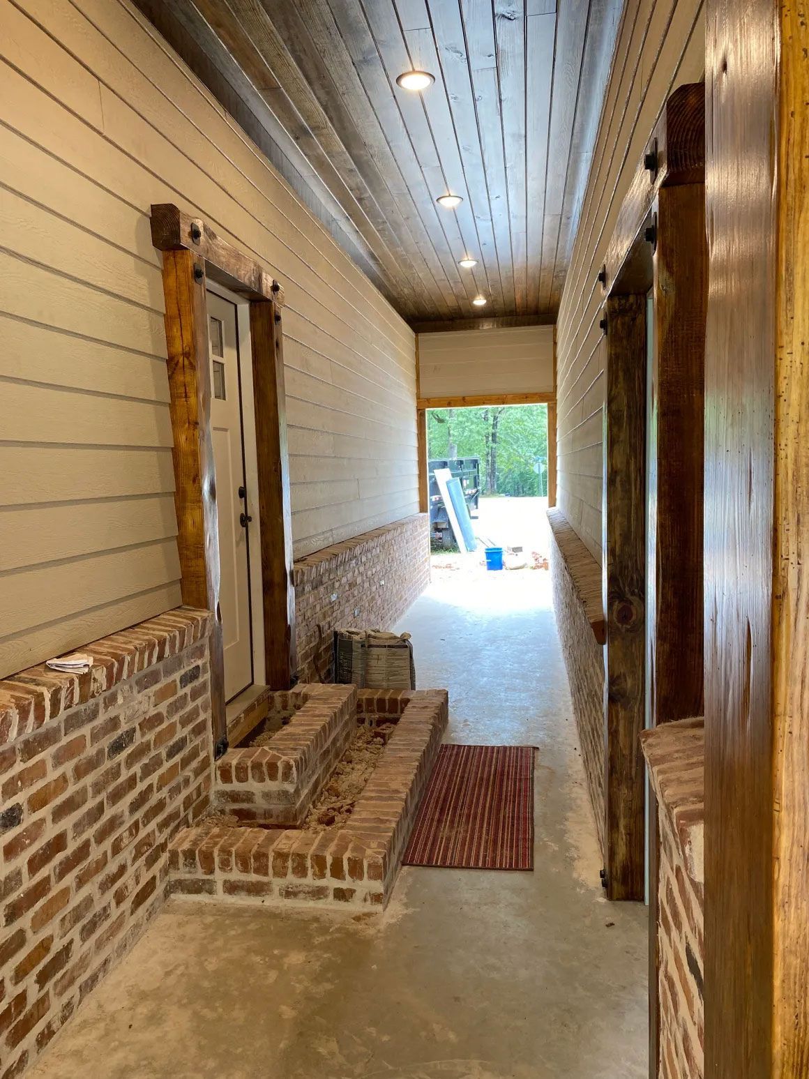 Narrow covered porch with brick steps, wooden siding, and a concrete floor leading to a bright yard.