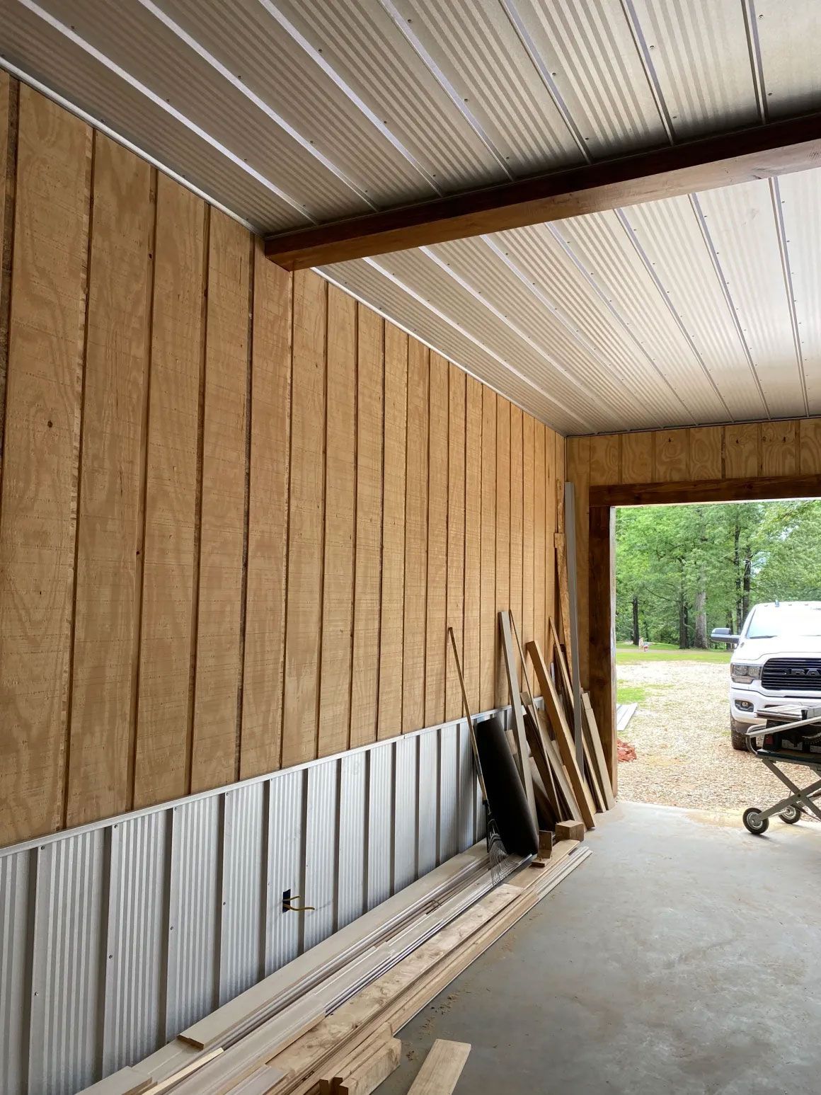 Covered wooden storage area with corrugated metal wall, lumber stacked along the side, and a driveway outside.