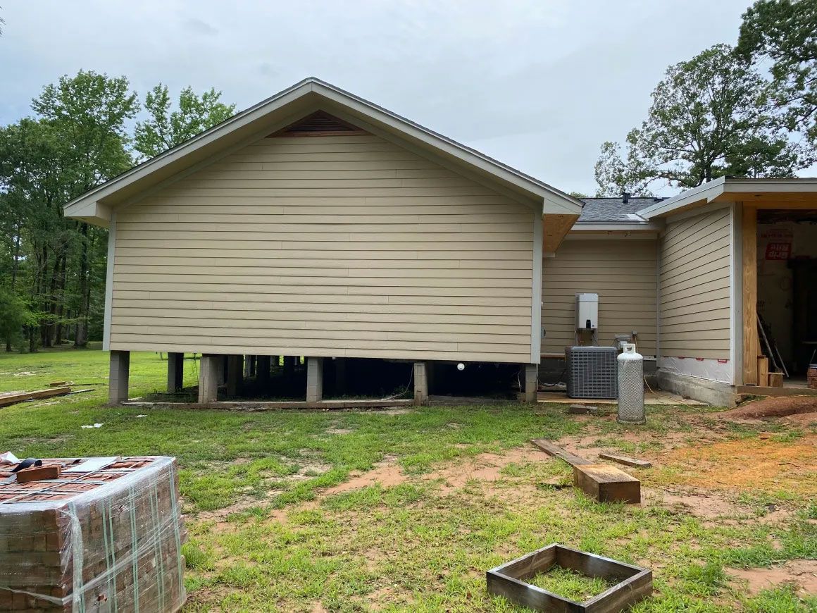 House exterior with raised siding and exposed foundation, set on a grassy lot with trees nearby