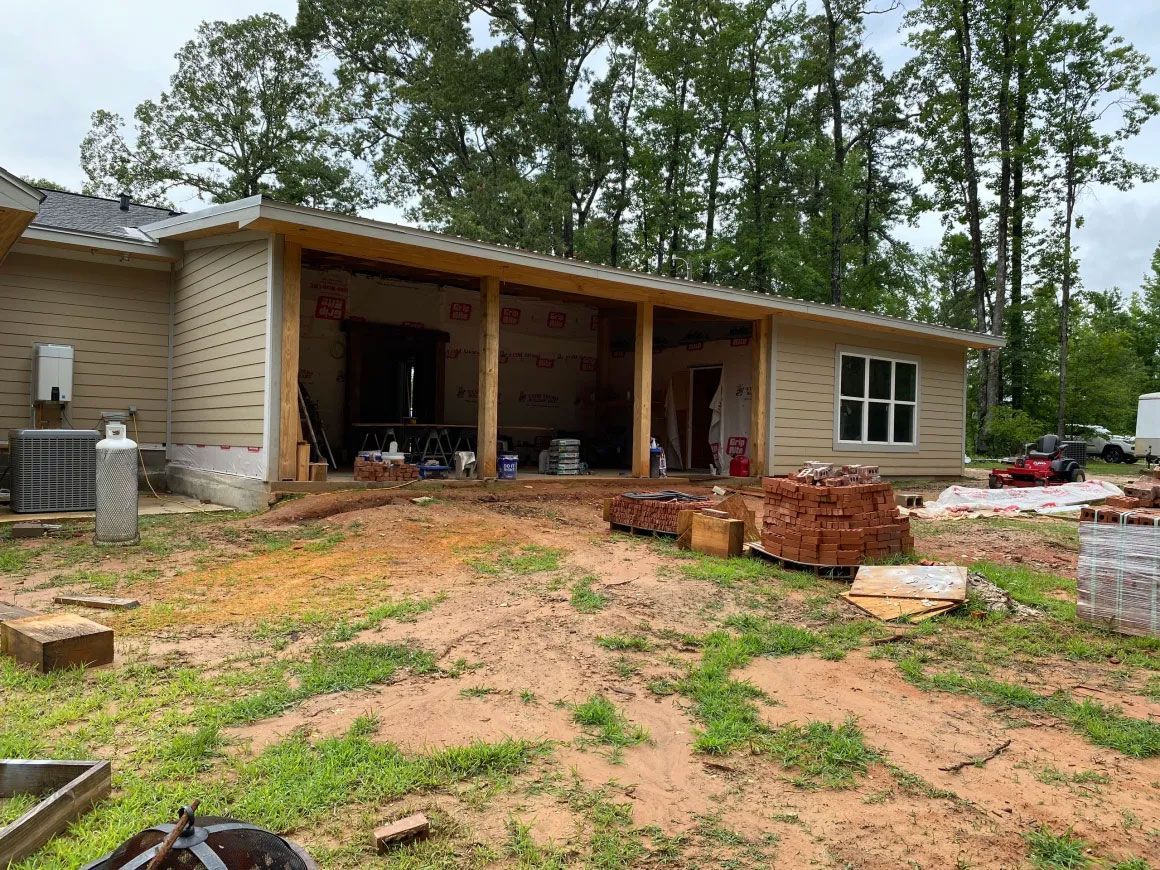 Partially finished house with open garage, brick, and dirt yard surrounded by trees