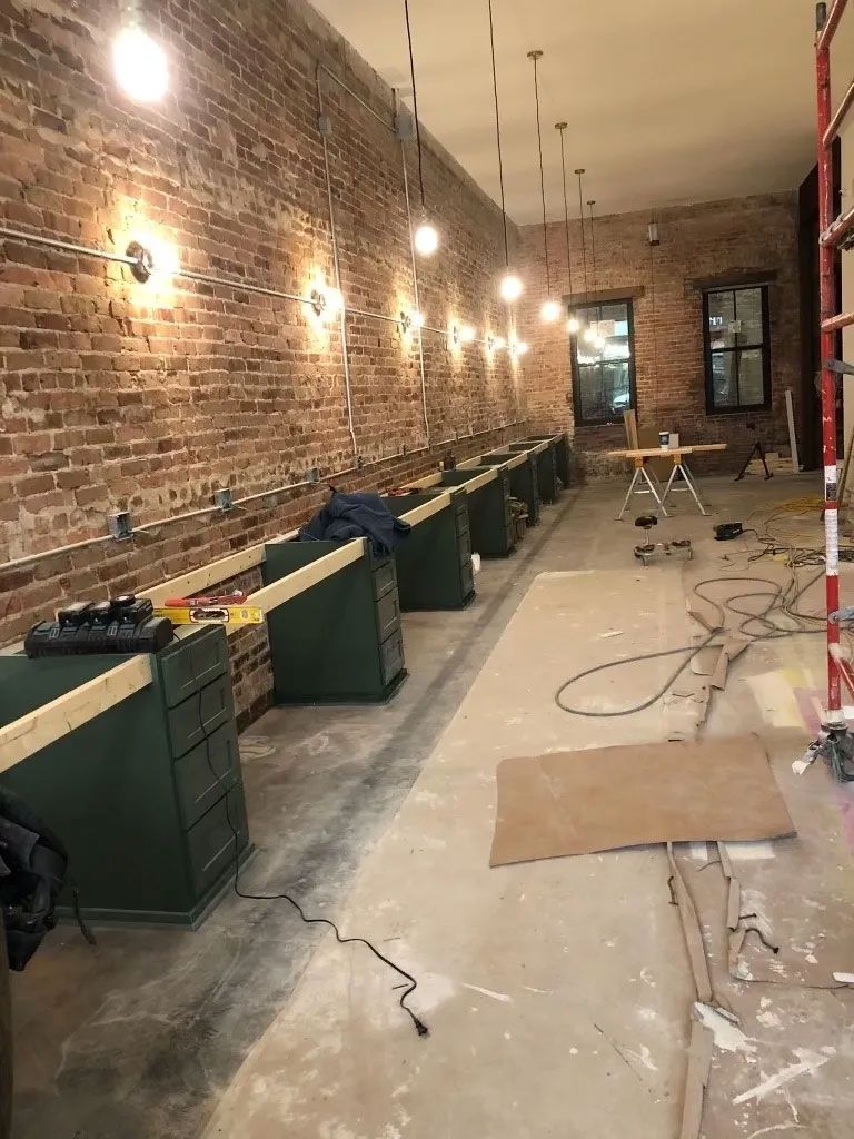 Narrow brick-walled room under construction with green counters, wall lights, and cables on a bare floor