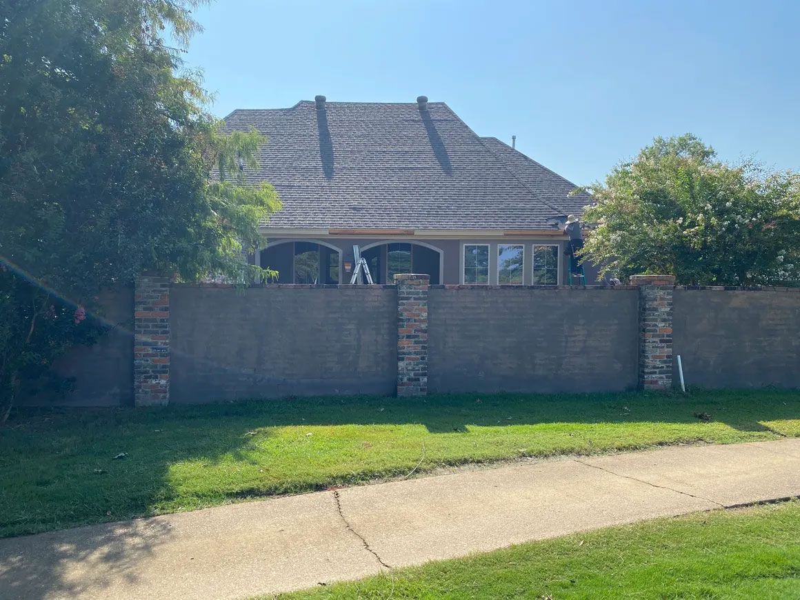 Single-story house behind a brick wall, with a grassy front yard and clear blue sky.