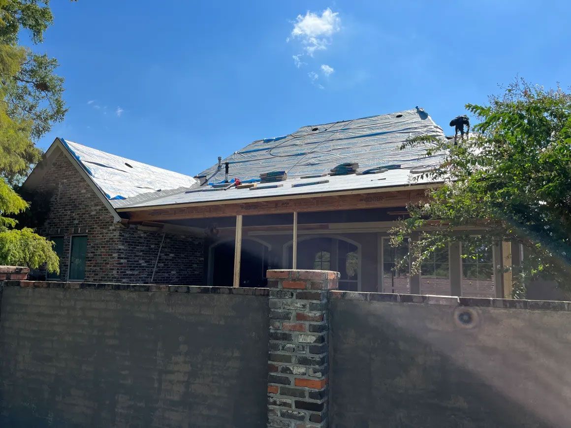 Partially damaged house roof with missing shingles, viewed over a brick wall under a blue sky.