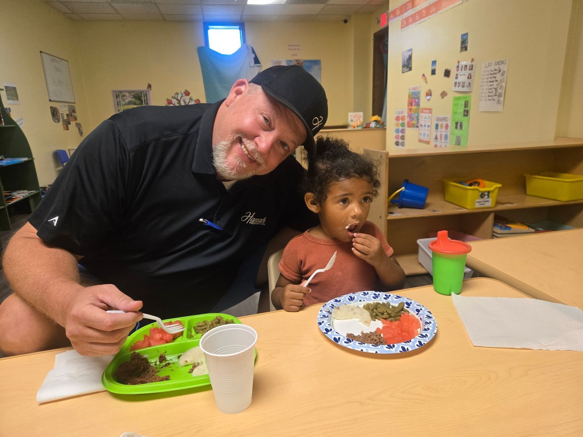 Man and child seated at a table eating, possibly in a classroom.
