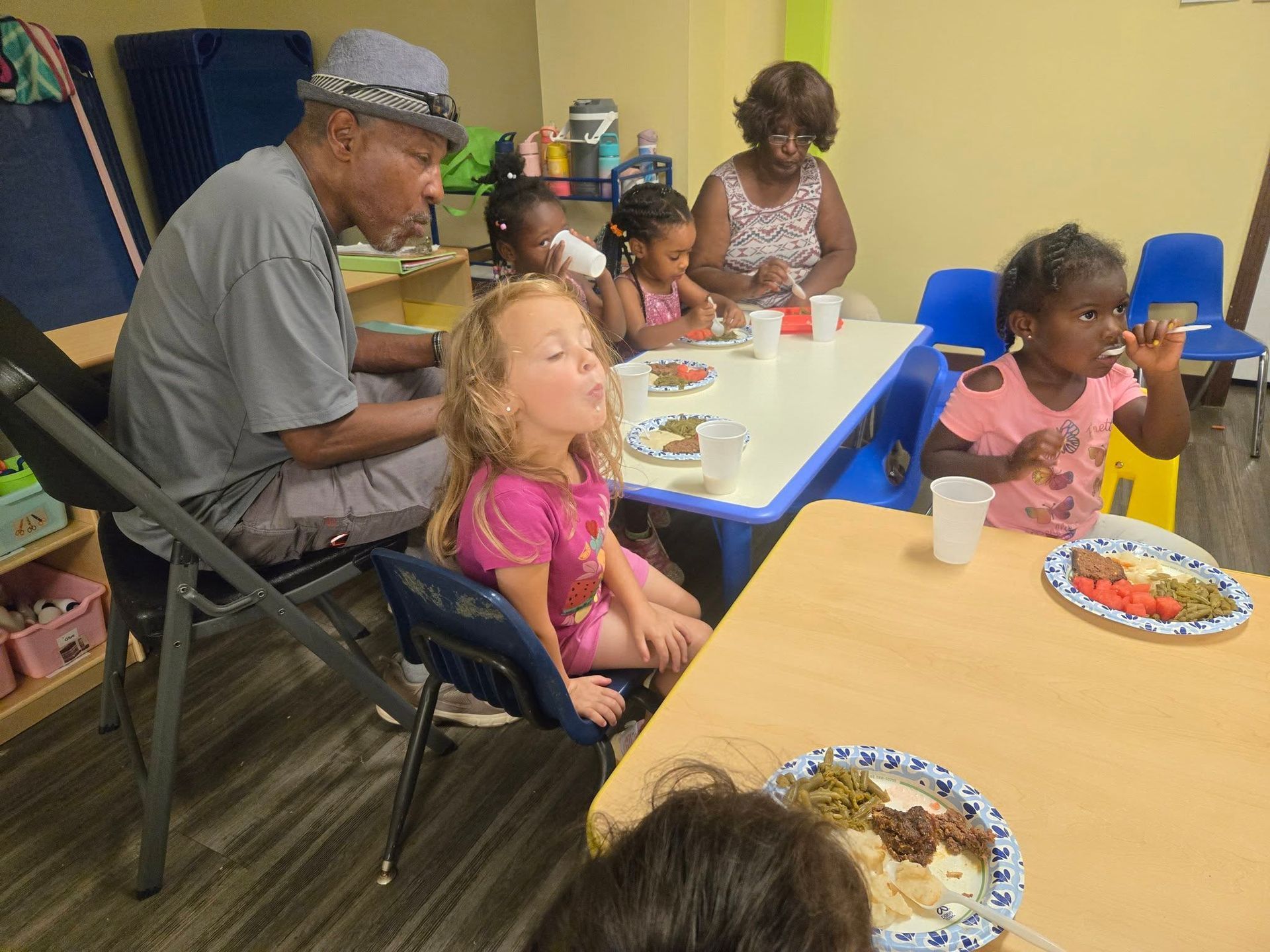 People eating at small tables in a room. An older man sits with children.
