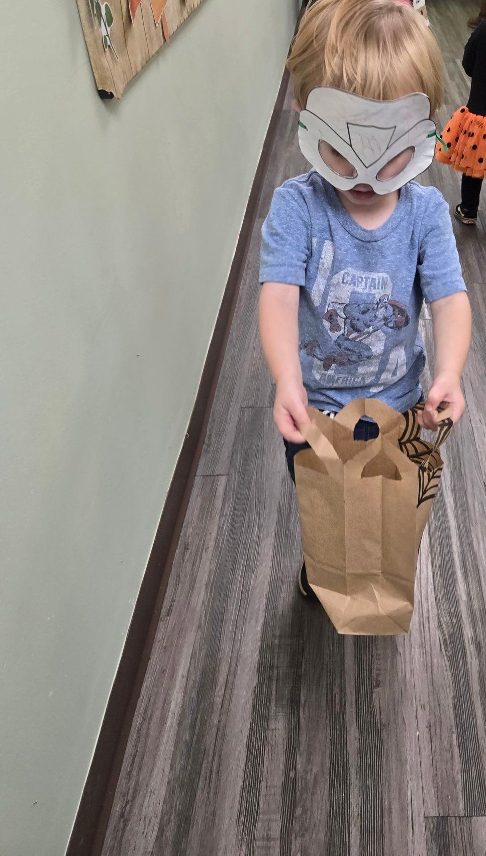 Child wearing a mask, holding a brown paper bag, walking down a hallway.