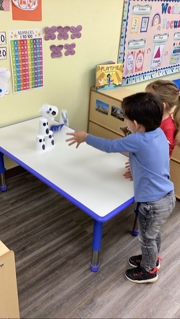 Two children playing a game in a classroom, knocking over stacked cups on a white table.