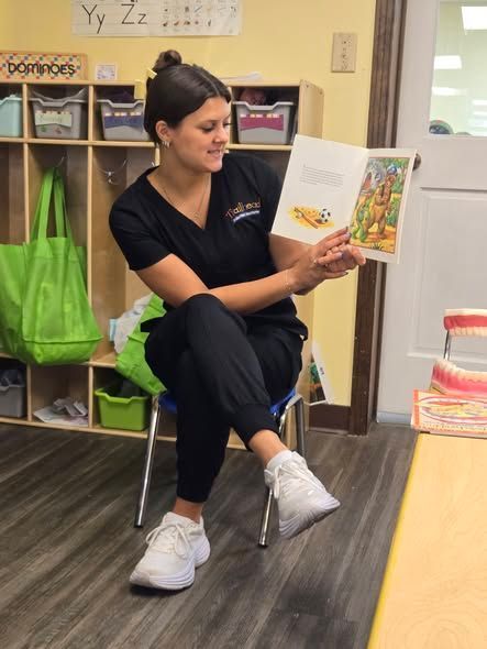 Woman in black scrubs reading a book to children in a classroom.