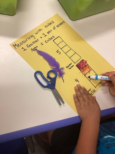 Child measuring with cubes on yellow paper next to feather and scissors.