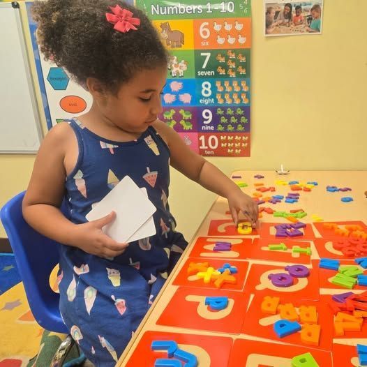Girl at a table with letter tiles, arranging them on red cards. Numbers chart in background.